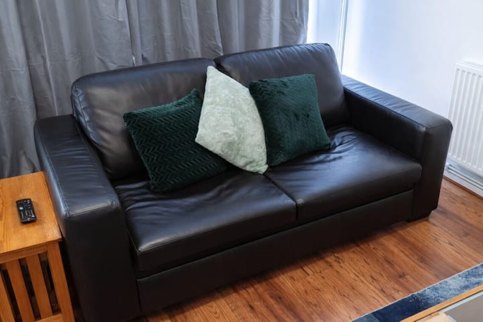 A dark brown leather sofa with two pillows, a remote control on a wooden side table, and a window with curtains in the background.