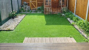 Ein Garten mit Kunstgrashalm, einer Holzdiele, einem Holzzaun und einer kleinen Holzhütte mit Fenstern.