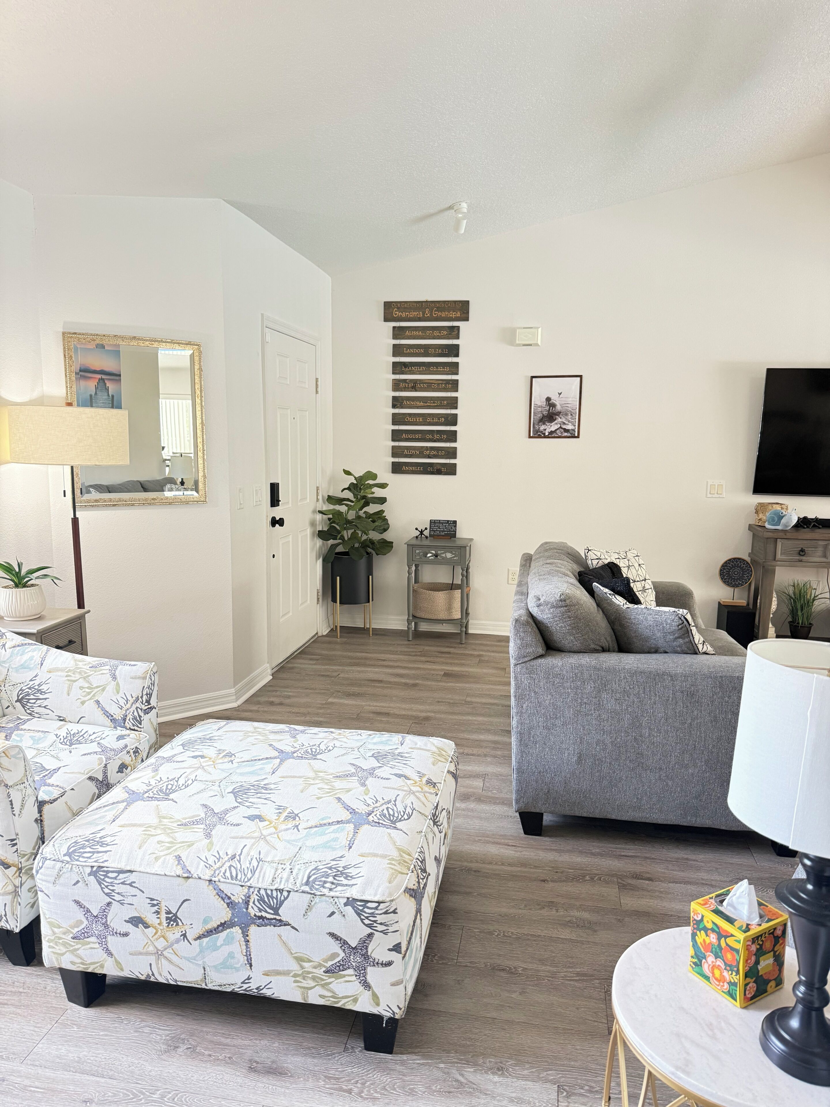Ein Wohnzimmer mit grauem Sofa, passendem Hocker, einem kleinen runden Tisch mit Lampe, einem Fernseher und einer Wandablage mit Dekoration.