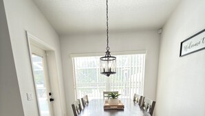A dining room with a large wooden table, chairs, a hanging light fixture, and a window with blinds. - Quaint, modern 3-bedroom house in charming, historic Inverness Florida. (Inverness)