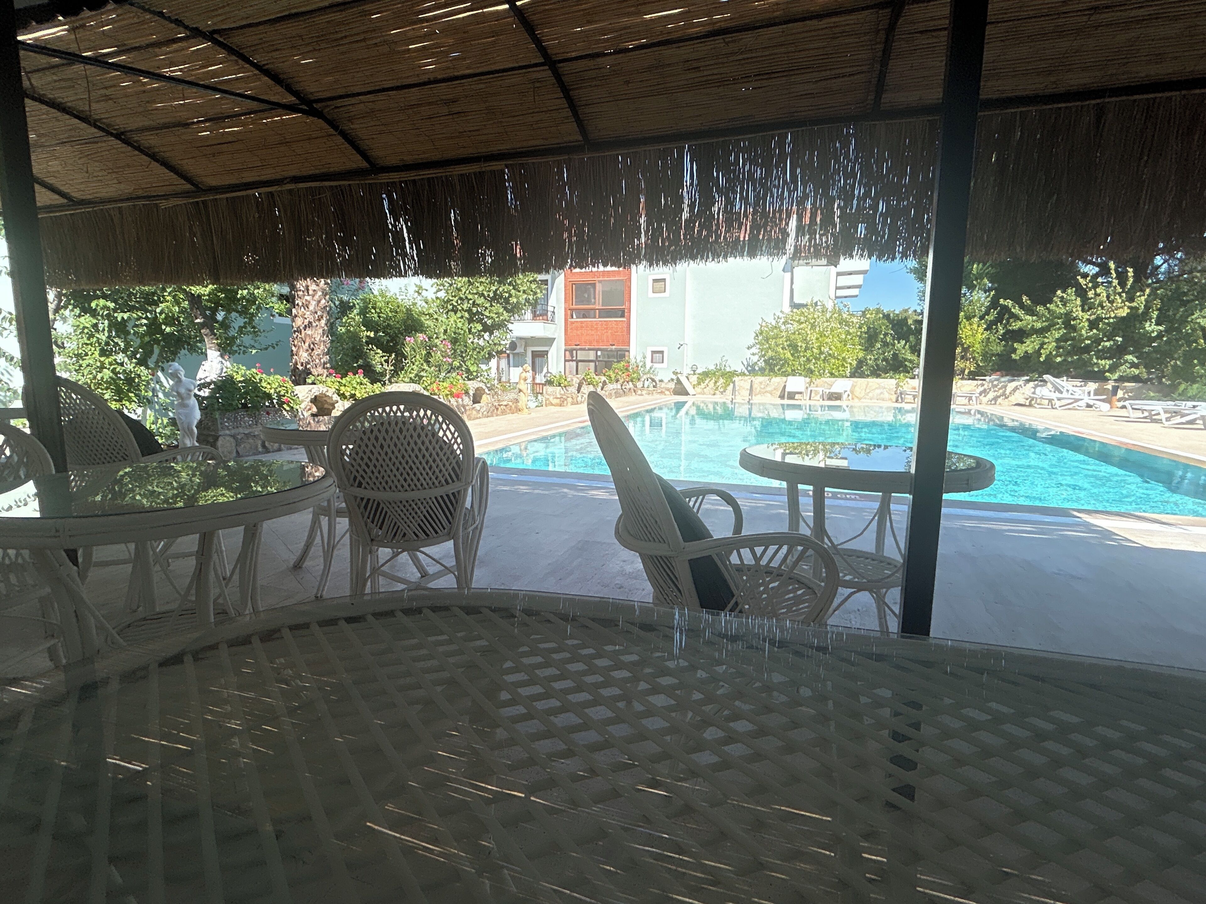Un área de estar techada al aire libre con piscina, sillas y vista a un edificio en el fondo.