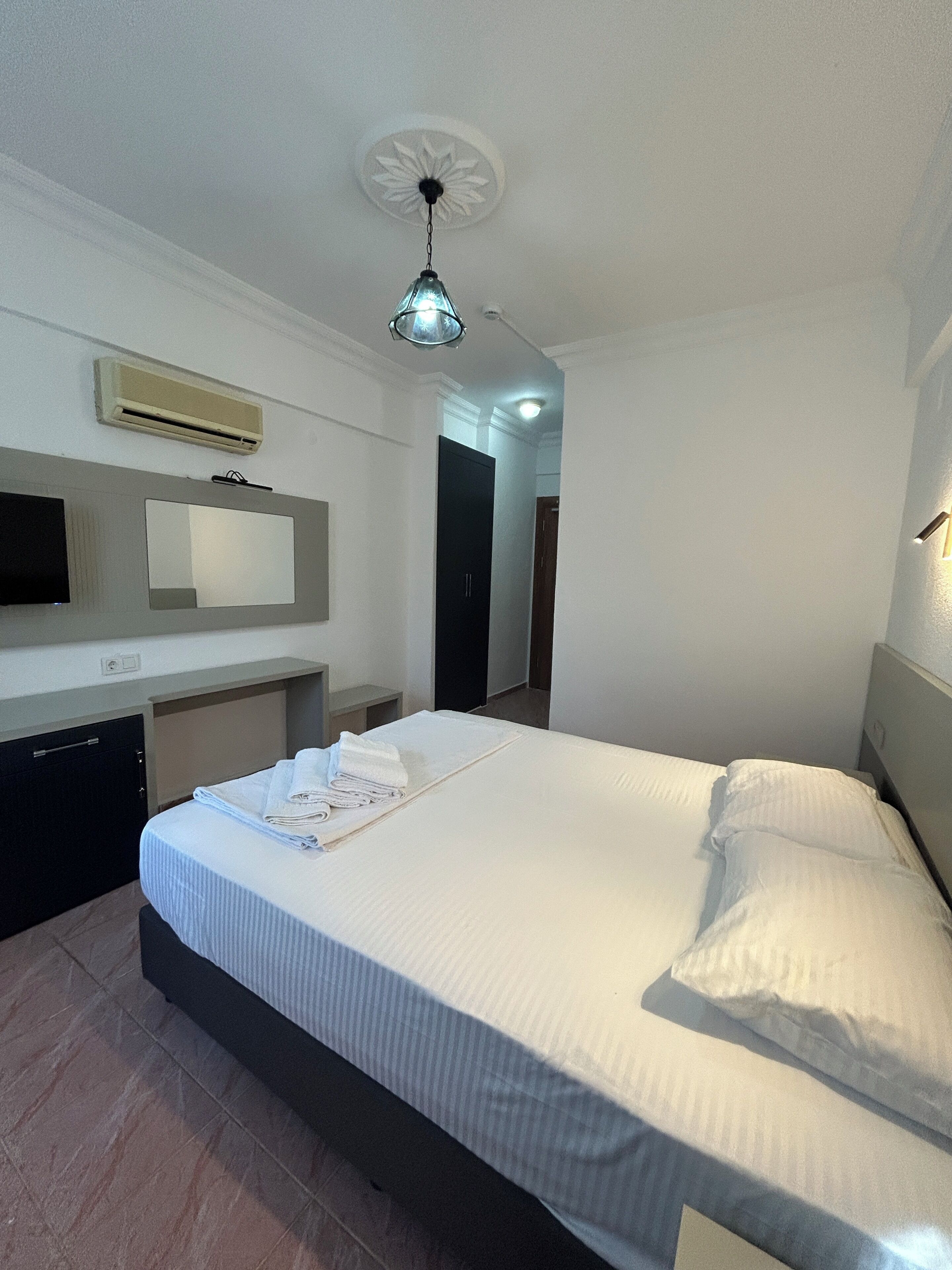 A hotel room with a bed, a desk with a chair, a flat-screen TV, an air conditioning unit, and a ceiling light fixture.