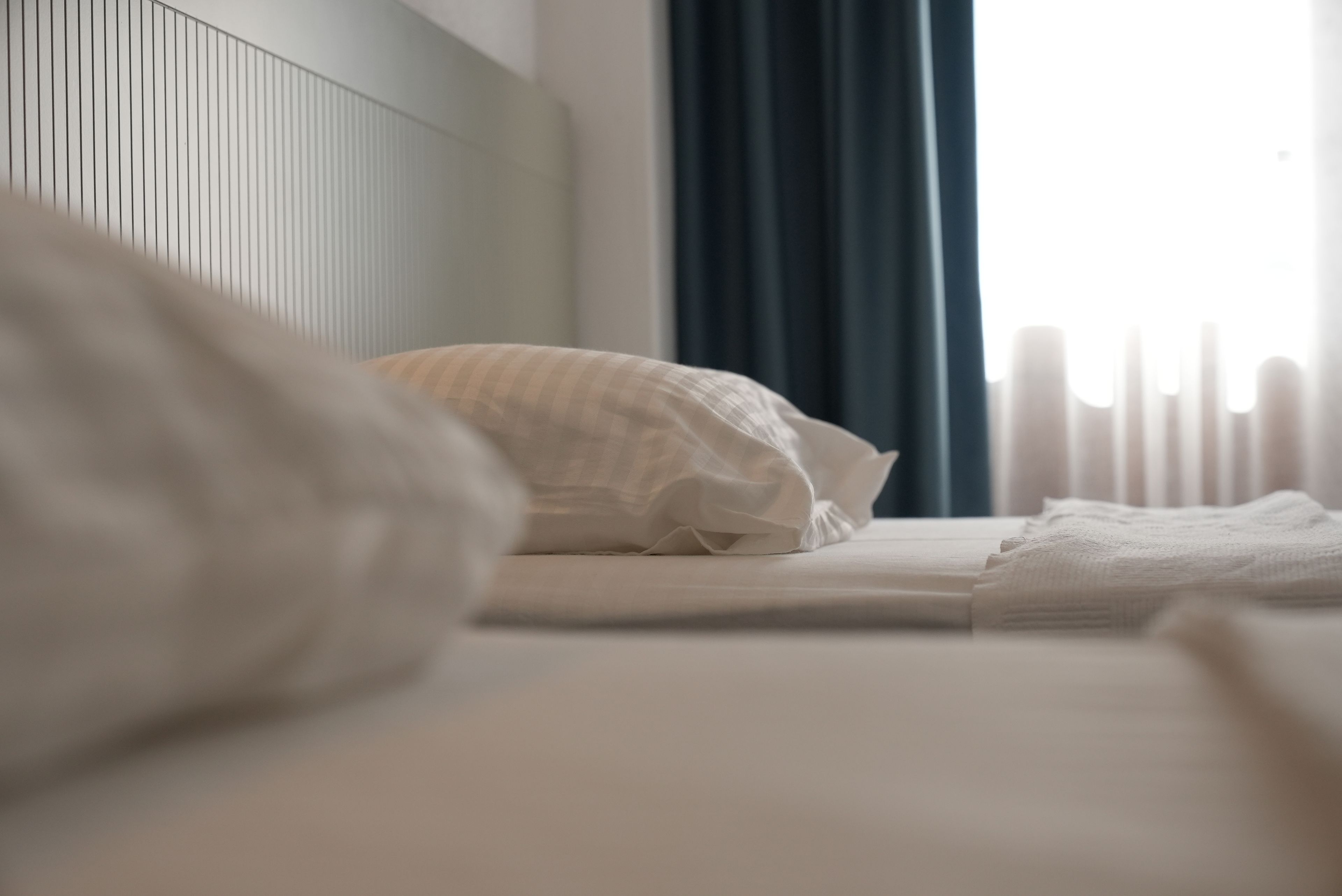 Una cama bien tendida con sábanas blancas y un cojín a rayas, una ventana con cortinas y un radiador.
