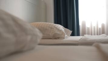 Una cama bien tendida con sábanas blancas y un cojín a rayas, una ventana con cortinas y un radiador.