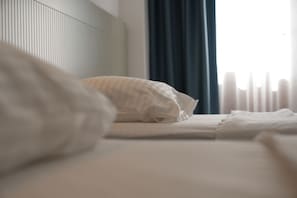 Una cama bien tendida con sábanas blancas y un cojín a rayas, una ventana con cortinas y un radiador.