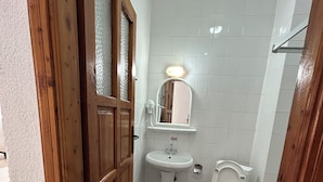 A bathroom with a wooden door, a white sink, a toilet, and a mirror with a light. - ÜNLÜ OTEL (Fethiye)