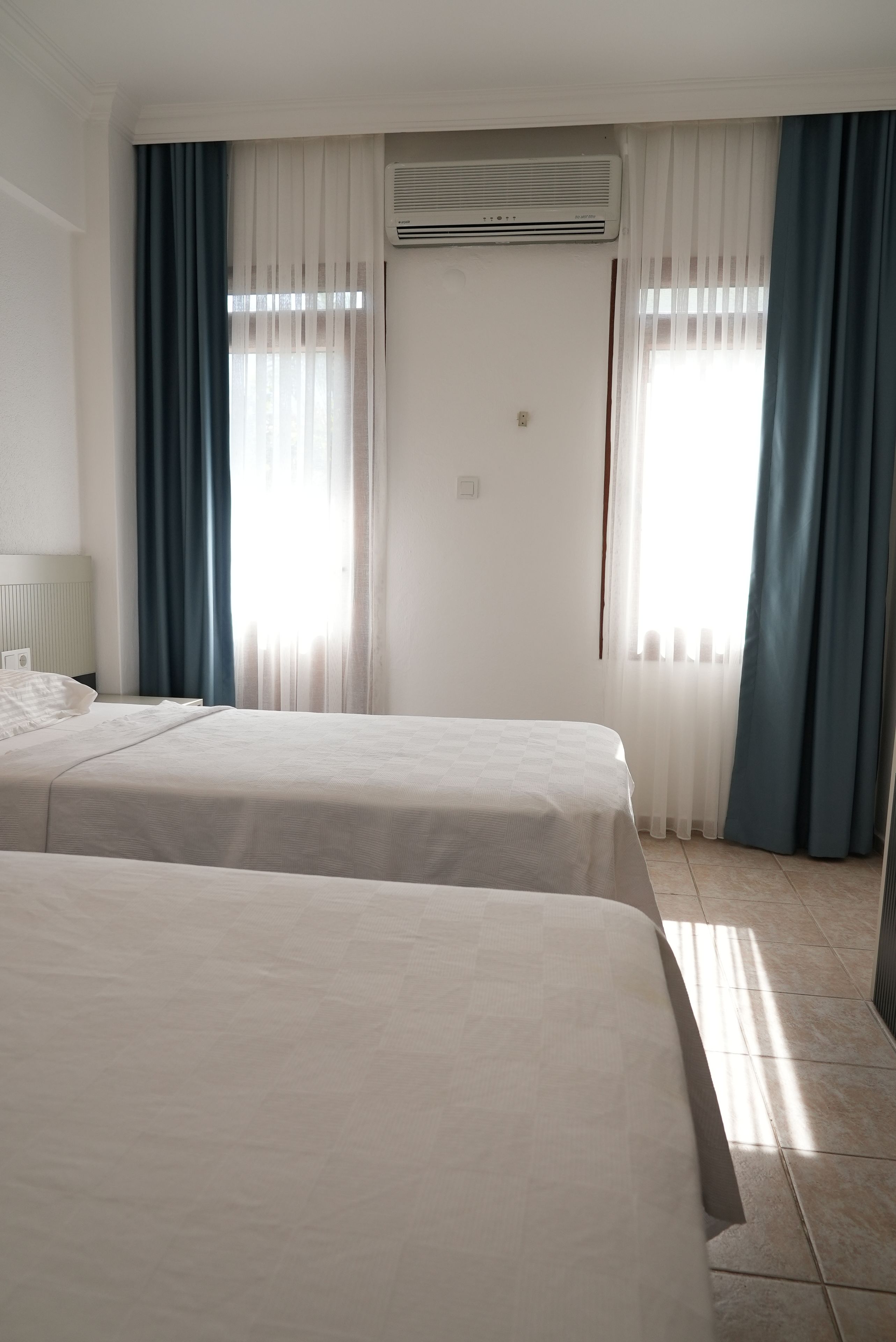 A hotel room with two beds, a window with curtains, and an air conditioning unit.