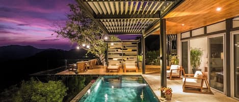 A modern outdoor pool area with a wooden deck, lounge chairs, and a view of the mountains at dusk.