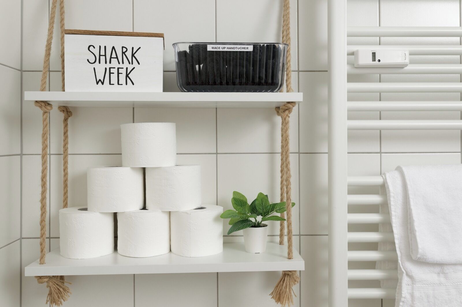 Et badeværelse med hvide fliser, en hylde med toiletpapir, en lille urtepotte, et skilt med teksten 'SHARK WEEK' og en toiletpapirholder hængt på væggen.