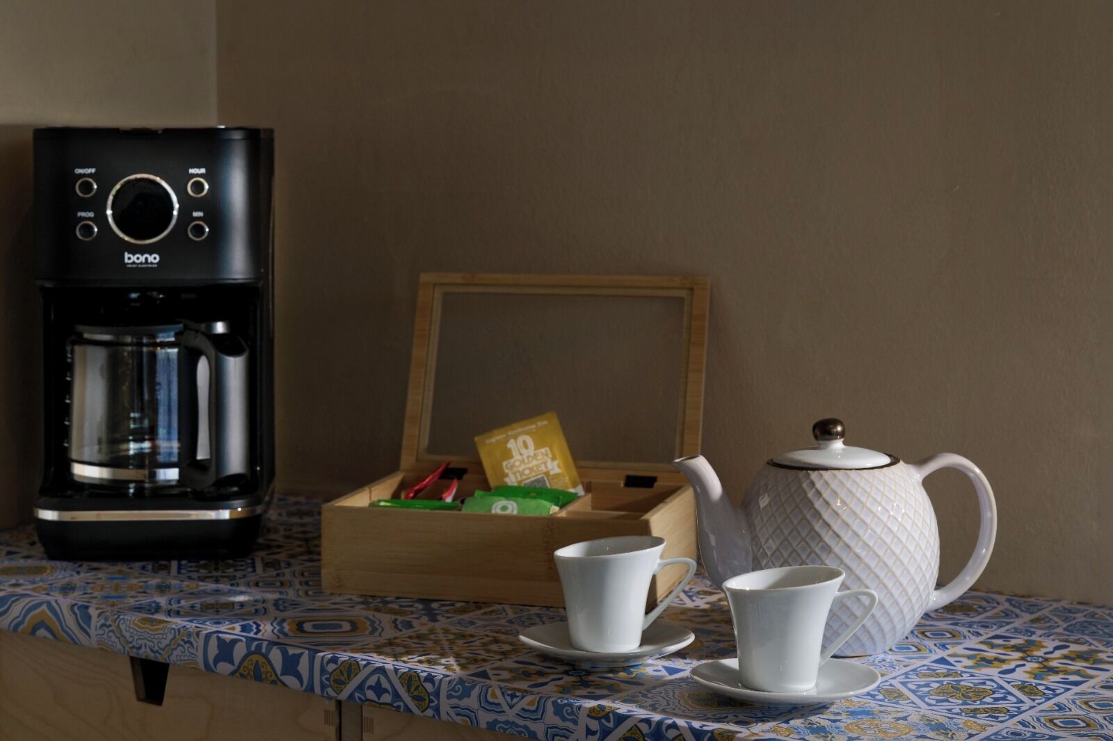 A coffee maker, a teapot, cups, and a box of tea bags on a table with a patterned tablecloth.
