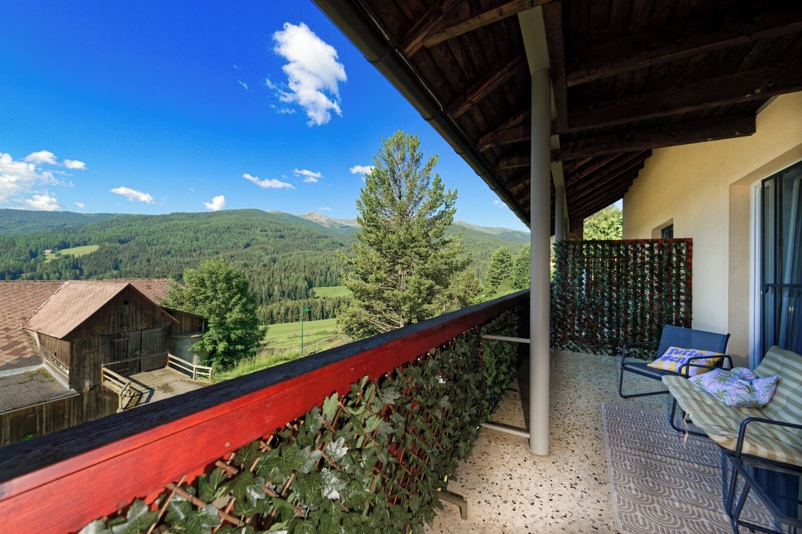 Ein Balkon mit Blick auf eine Berglandschaft, ein hölzernes Bauernhaus und eine überdachte Terrasse mit Stuhl und Tisch.