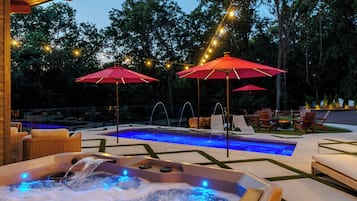Un jacuzzi con luz azul, rodeado de mobiliario exterior y sombrillas rojas, con una piscina y árboles al fondo.