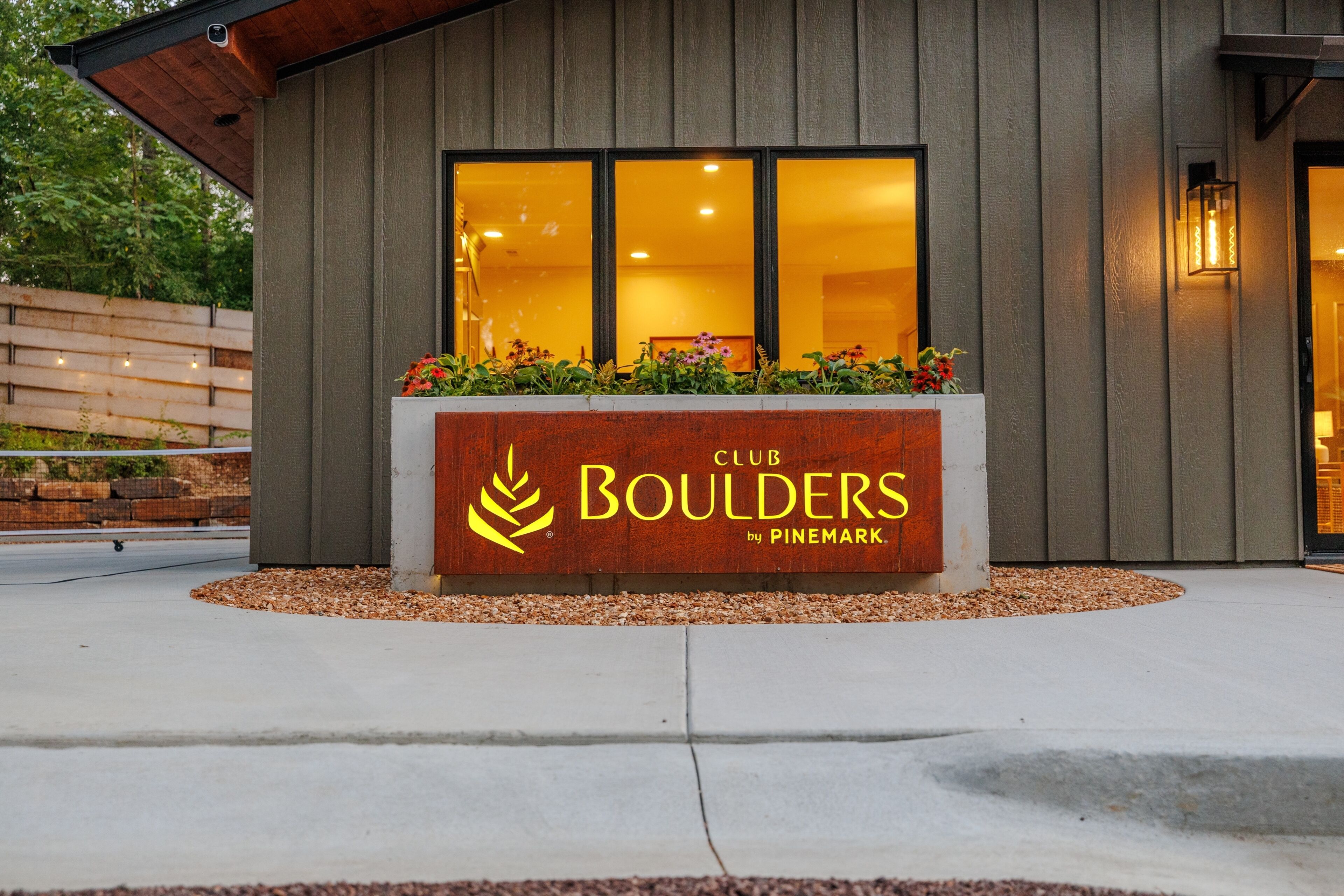 Een Club Boulders bord met een logo, bij de ingang van een gebouw met een houten gevel en een raam dat binnenverlichting weerspiegelt.