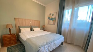 A bedroom with a bed, two bedside tables, a nightstand, a rug, a window with curtains, and a wall art piece. - Ground-floor apartment with spacious terraces, 15 minutes from the marina and Falésia Beach (Quarteira)