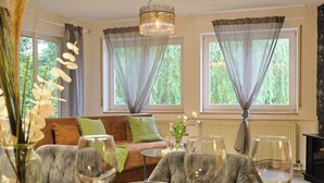 Un salon avec un canapé, une table à manger avec des verres à vin, un lustre et de grandes fenêtres avec des rideaux.