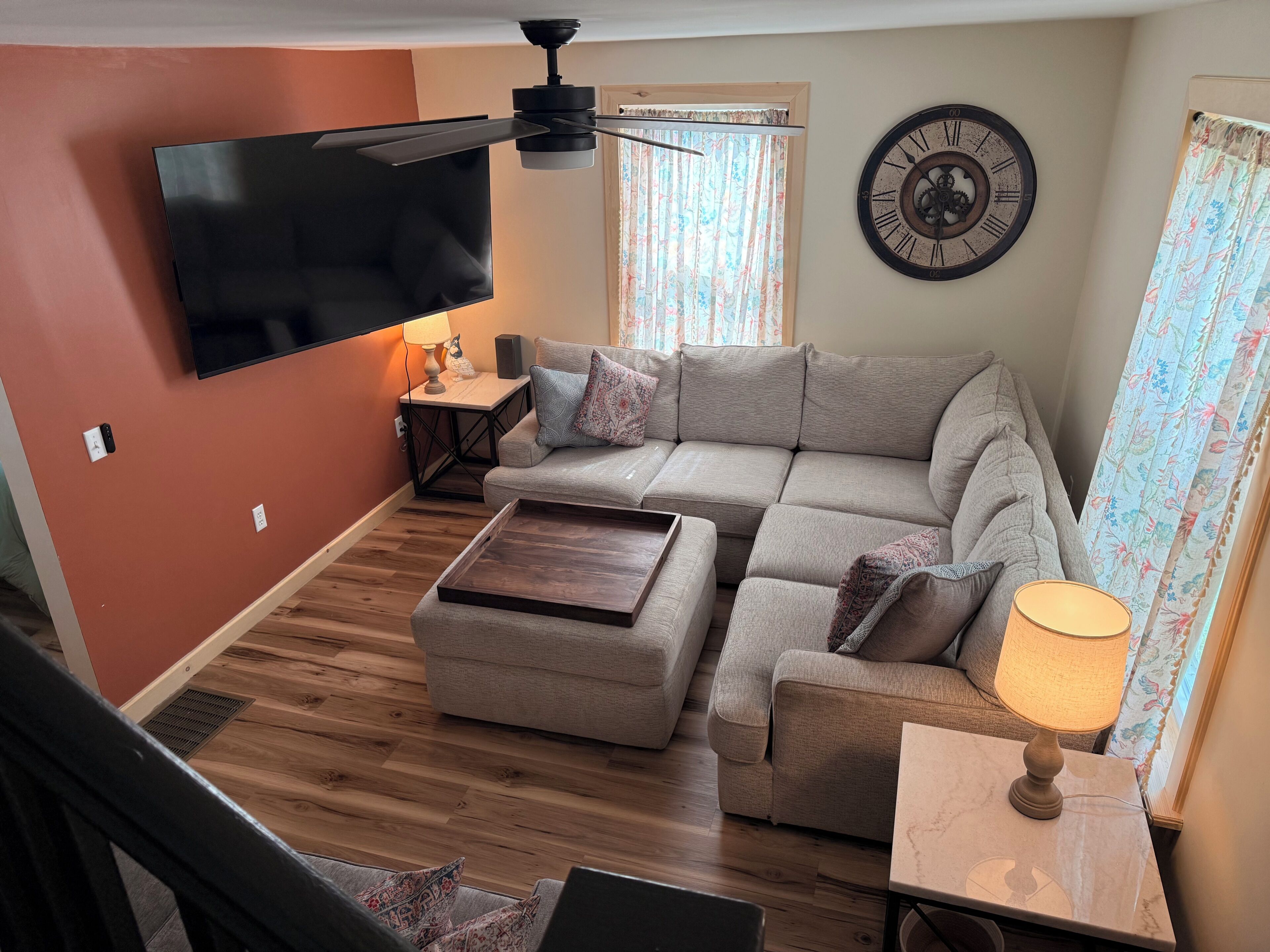 Un salon comprenant un canapé d’angle, un téléviseur à écran plat fixé au mur, un ventilateur de plafond, une horloge et une fenêtre avec des rideaux.