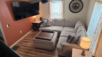 Un salon comprenant un canapé d’angle, un téléviseur à écran plat fixé au mur, un ventilateur de plafond, une horloge et une fenêtre avec des rideaux.