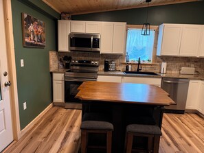 A kitchen with white cabinets, stainless steel appliances, a wooden table, and two bar stools. - Serene 3-bedroom house with AC in Alexandria Bay (Alexandria Bay)