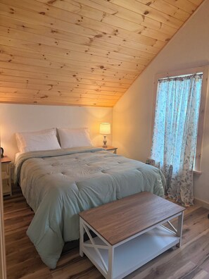 A bedroom with a wooden ceiling, a bed with a light-colored duvet, a bedside table with a lamp, and a window with floral curtains. - Serene 3-bedroom house with AC in Alexandria Bay (Alexandria Bay)