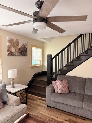 A living room with a gray sofa, a table with a lamp, a ceiling fan, and a staircase with black railings. - Serene 3-bedroom house with AC in Alexandria Bay (Alexandria Bay)