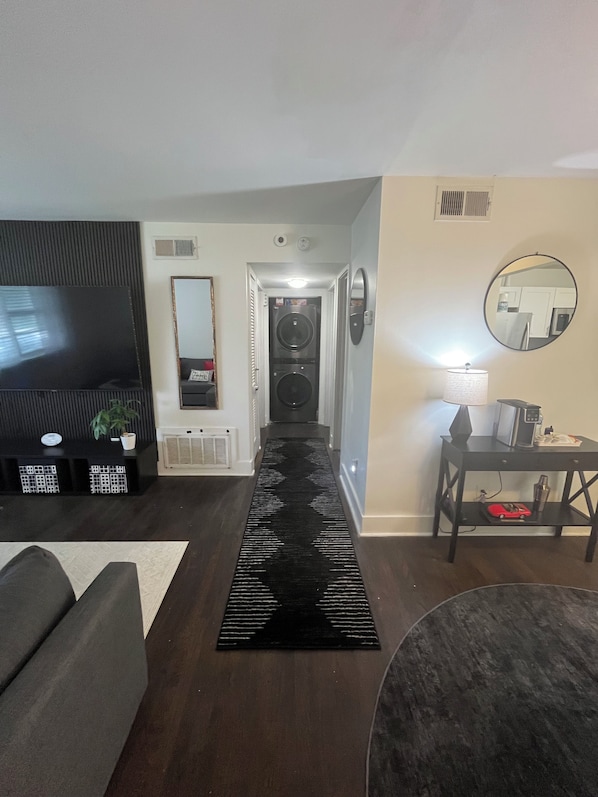 A living room with a black rug, a sofa, a TV, a washing machine, a mirror, and a table with a lamp.