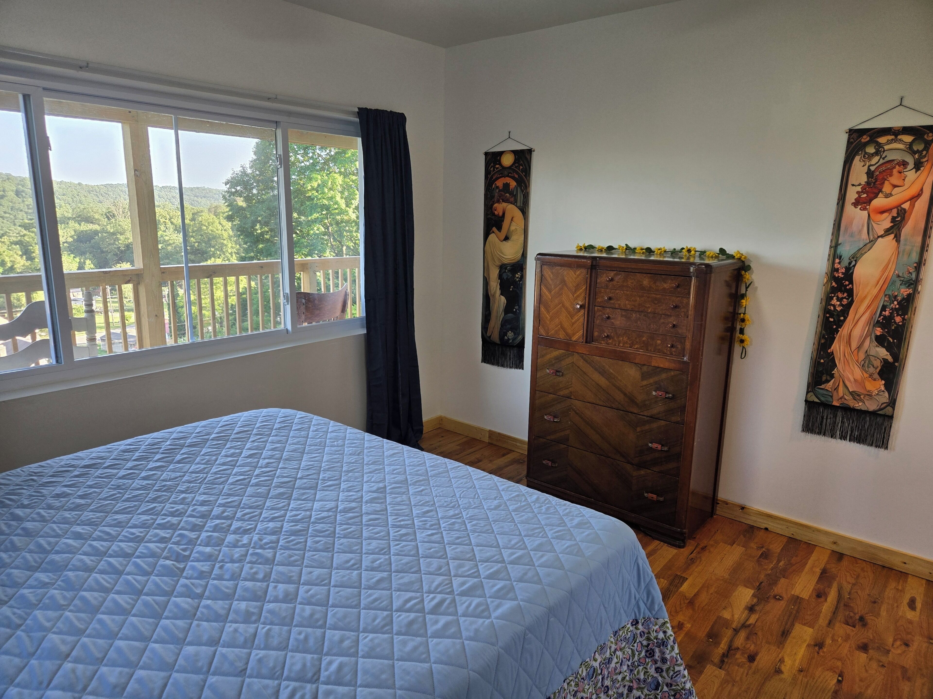 A bedroom with a bed, a wooden dresser, a window with a view, and two wall hangings.