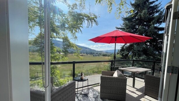 Ein Balkon mit Korbmöbeln, einem roten Sonnenschirm und Blick auf eine grüne Landschaft.