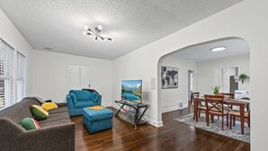 A living room with a sofa, ottoman, and a TV stand. There is a dining area with a table and chairs. - Independence House Near GEHA Field & Fenced Yard (Independence)