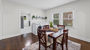 A dining area with a wooden table and chairs, a built-in oven, a washing machine, and a window with blinds. - Independence House Near GEHA Field & Fenced Yard (Independence)