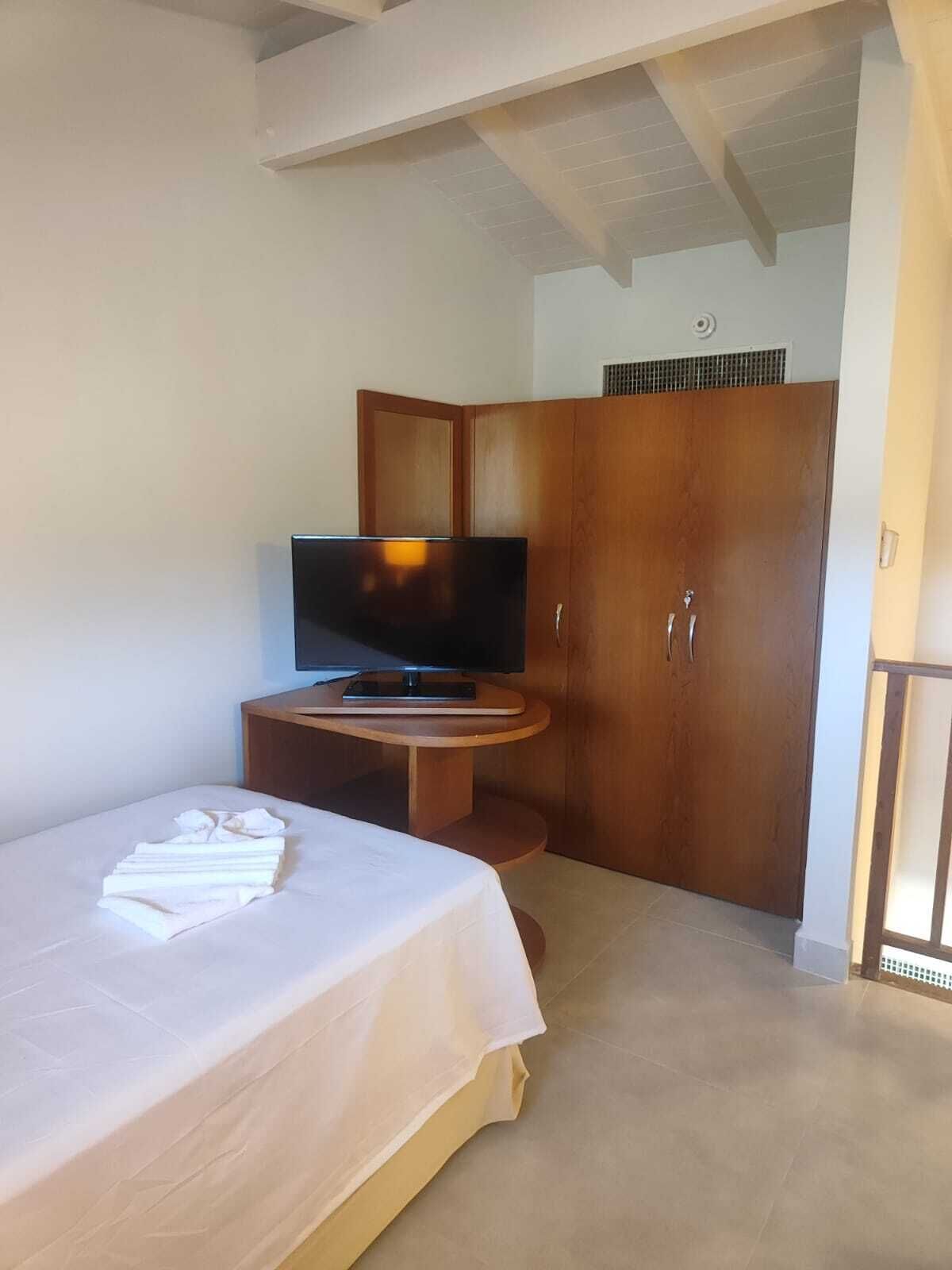 Habitación de hotel con cama, televisor sobre un pedestal de madera, armario y una mesita.