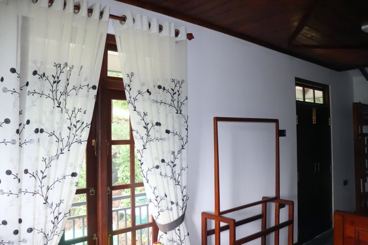 A room with a wooden door, a window with a view of greenery, and a white curtain with a floral pattern.
