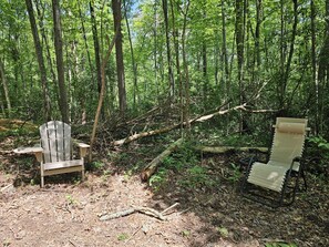 A forested area with two wooden chairs and a table, surrounded by trees and natural ground cover.