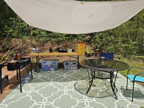 An outdoor camping setup with a table, chairs, a grill, and storage containers.