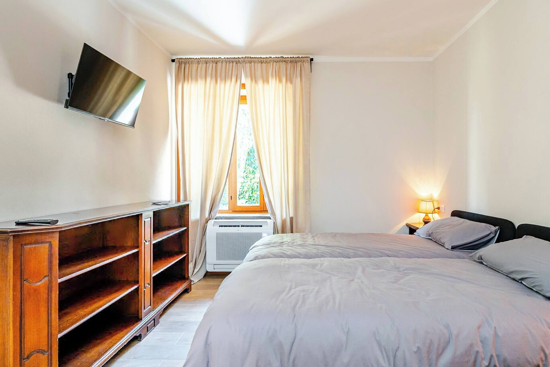 Quarto com cama, televisão na parede, escrivaninha com cadeira e janela com cortinas.