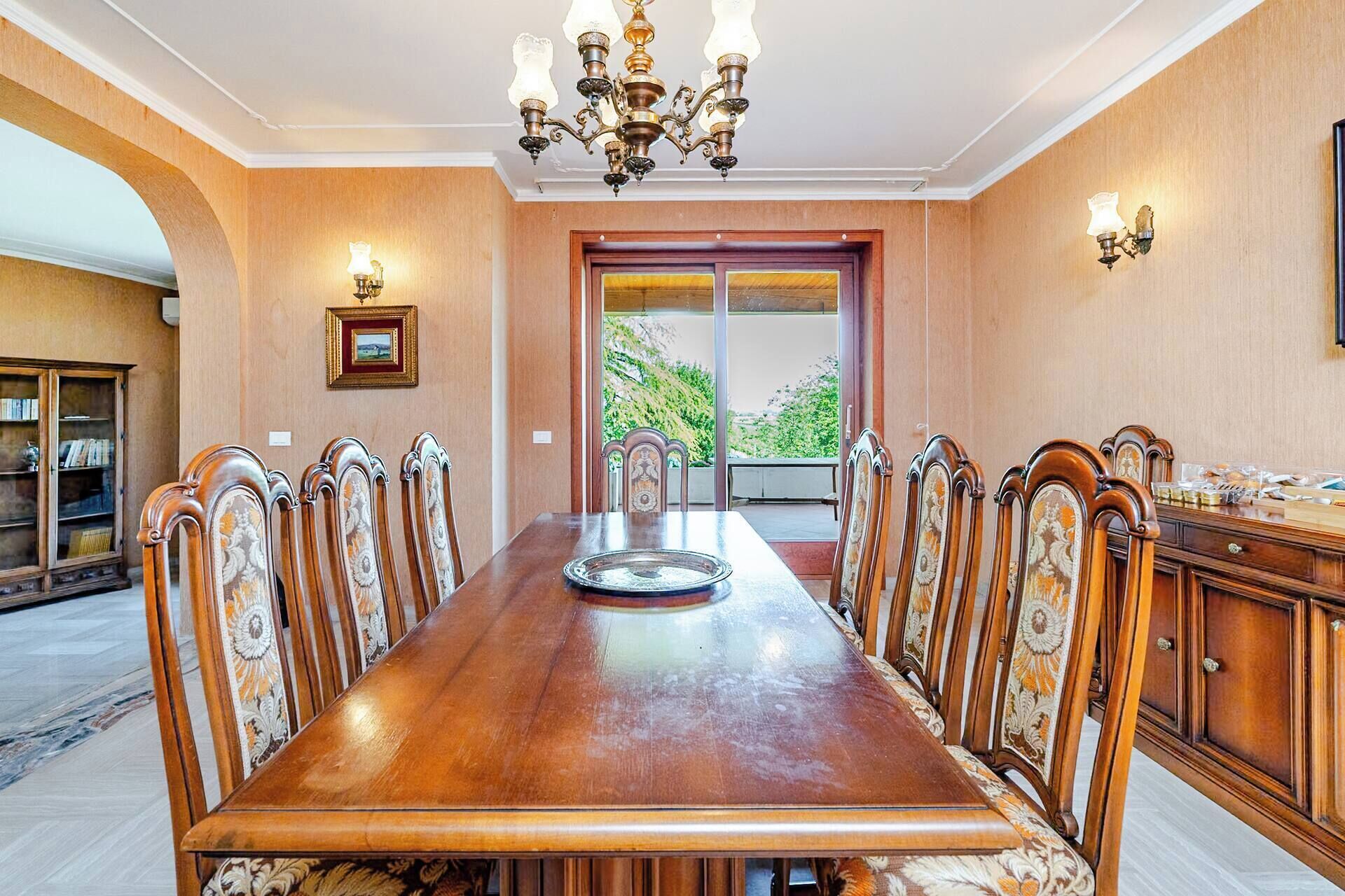 Una sala da pranzo con un lungo tavolo di legno, sedie decorate, un lampadario e vista attraverso una porta vetrata.