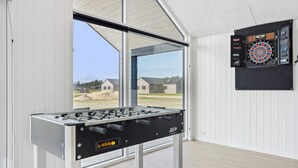 A foosball table in a room with large windows, a dartboard, and a view of houses outside. - 5 Star Holiday Home in Norre Nebel (Norre Nebel)