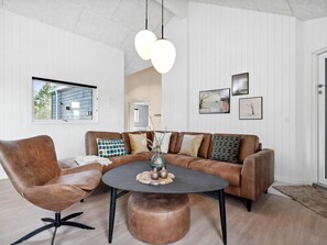 A modern living room with a brown leather sofa, a round coffee table, and a brown leather armchair. There is a window with a view of a house outside. - 5 Star Holiday Home in Norre Nebel (Norre Nebel)