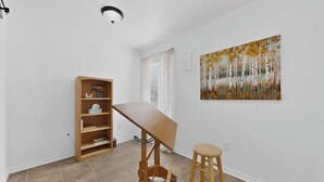 Una habitación con un escritorio de madera, un taburete, una estantería y un cuadro en la pared.