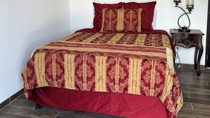 A bedroom with a large bed, a decorative wall hanging, a small table with two lamps, and a tiled floor.