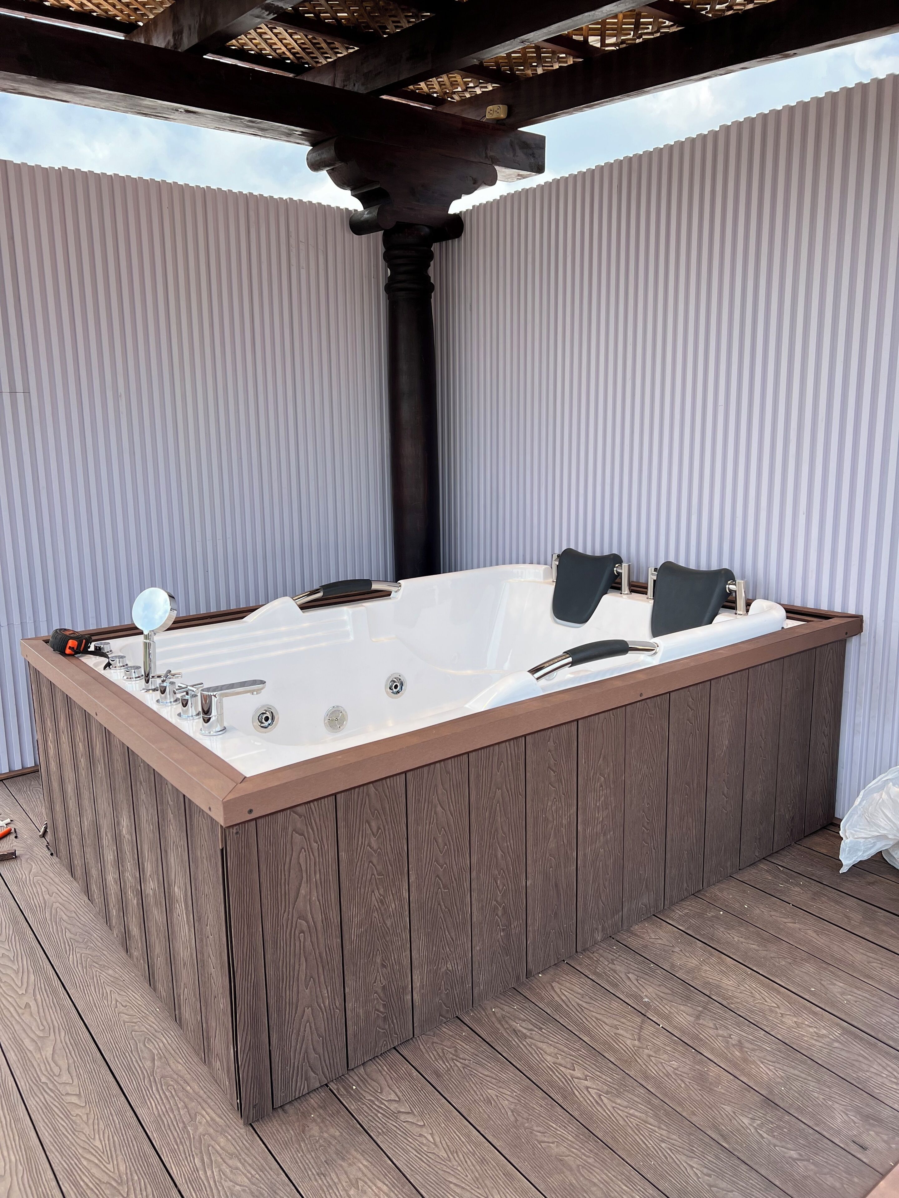 A hot tub with a wooden frame, a faucet, and a control panel, set on a wooden deck with a pergola overhead.
