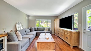 Ein Wohnzimmer mit einem Ecksofa, einem Couchtisch, Holzelementen, einem Fernseher und großen Fenstern.