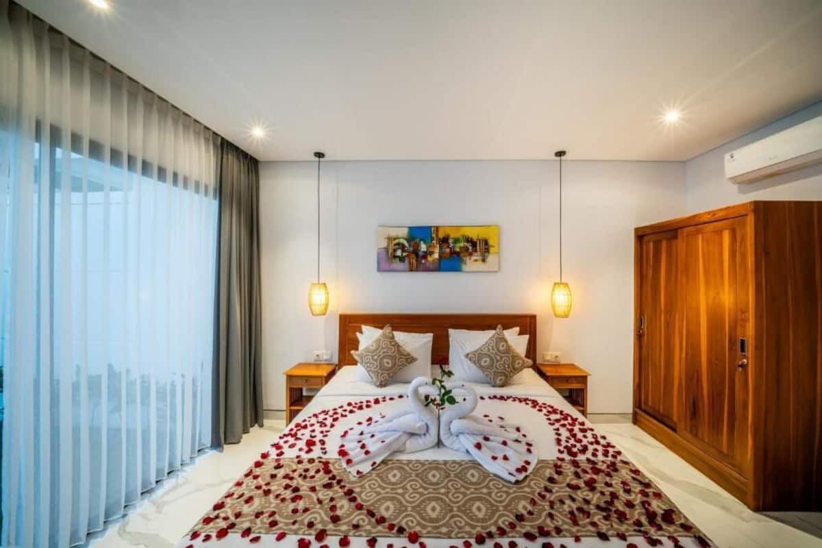 Une chambre d’hôtel avec un lit, deux tables de chevet, une armoire en bois, un tableau au mur et une grande fenêtre avec des rideaux.