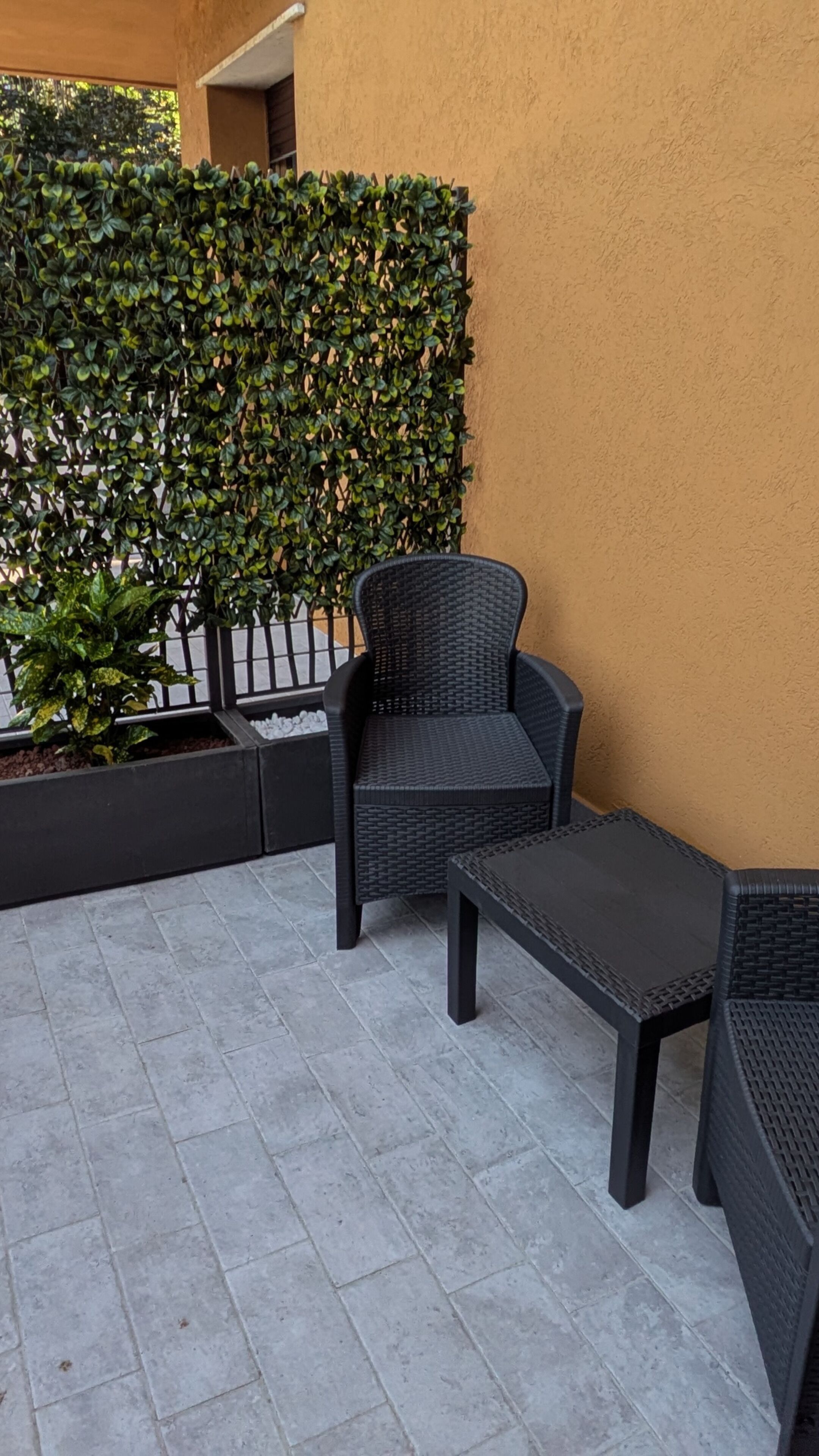 Un espacio al aire libre con una silla y una mesa, un seto y un piso de baldosas.