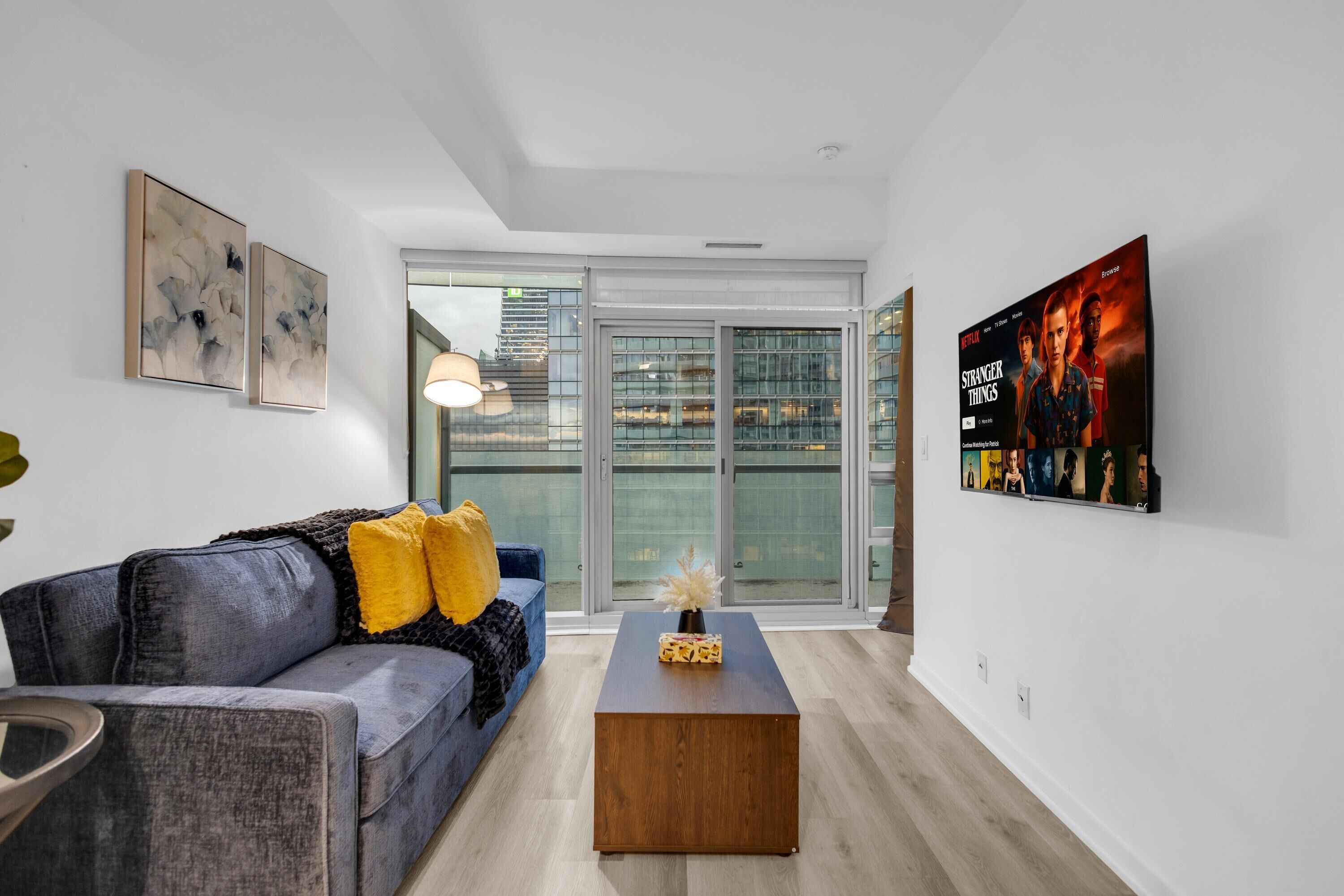 A modern living room with a grey sofa, a wooden coffee table, and a large window with a view of buildings.