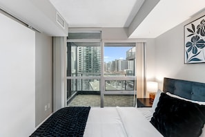 A hotel room with a bed, a nightstand, a lamp, a large window with a city view, and a door leading to a balcony. - Queen Verity Suites (Toronto)