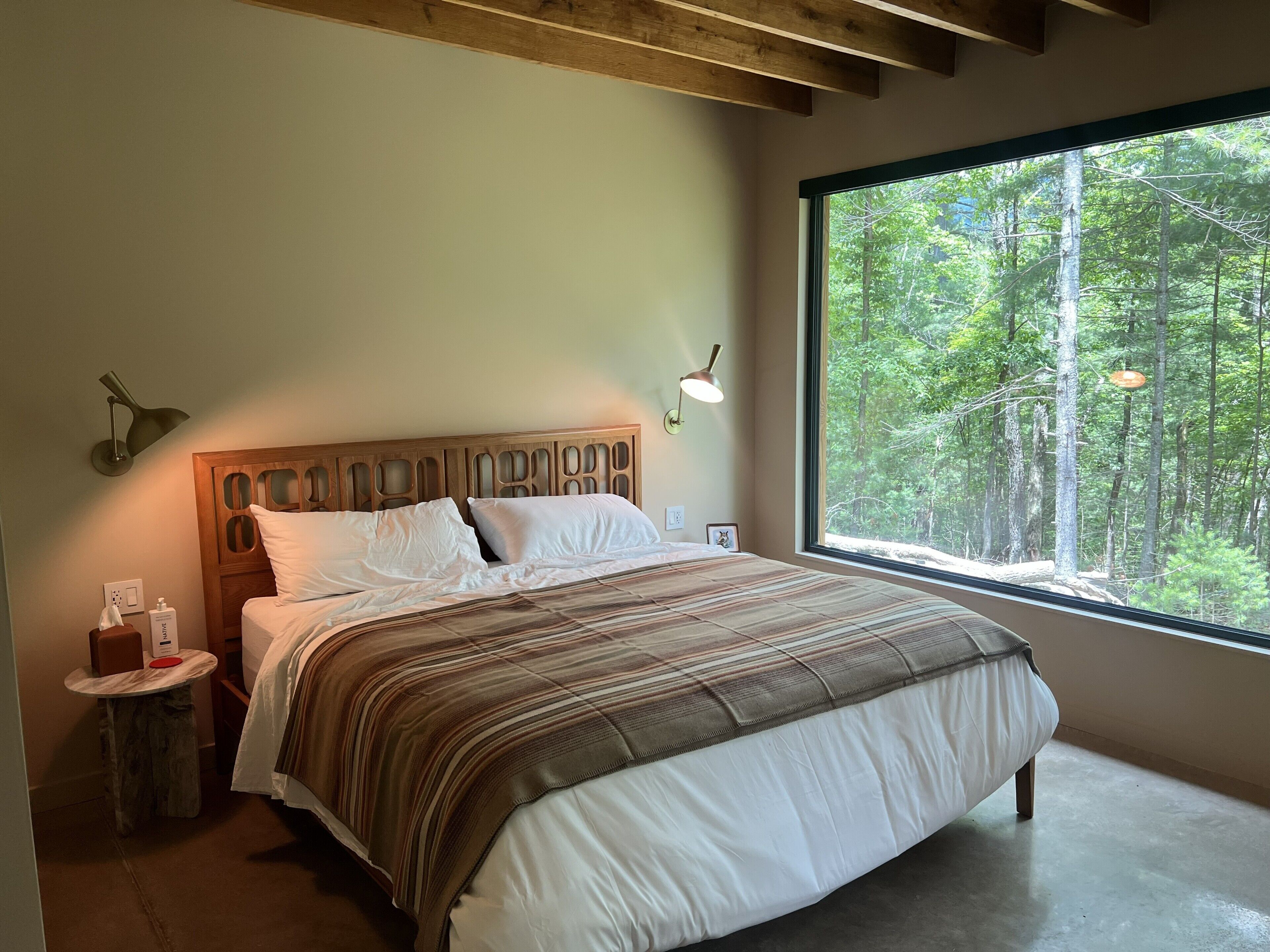 Une chambre avec une grande fenêtre, une tête de lit en bois, une table de chevet et une lampe.