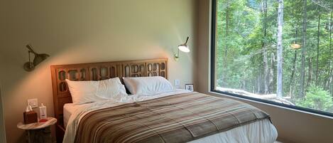 A bedroom with a large window, a wooden headboard, a bedside table, and a lamp.