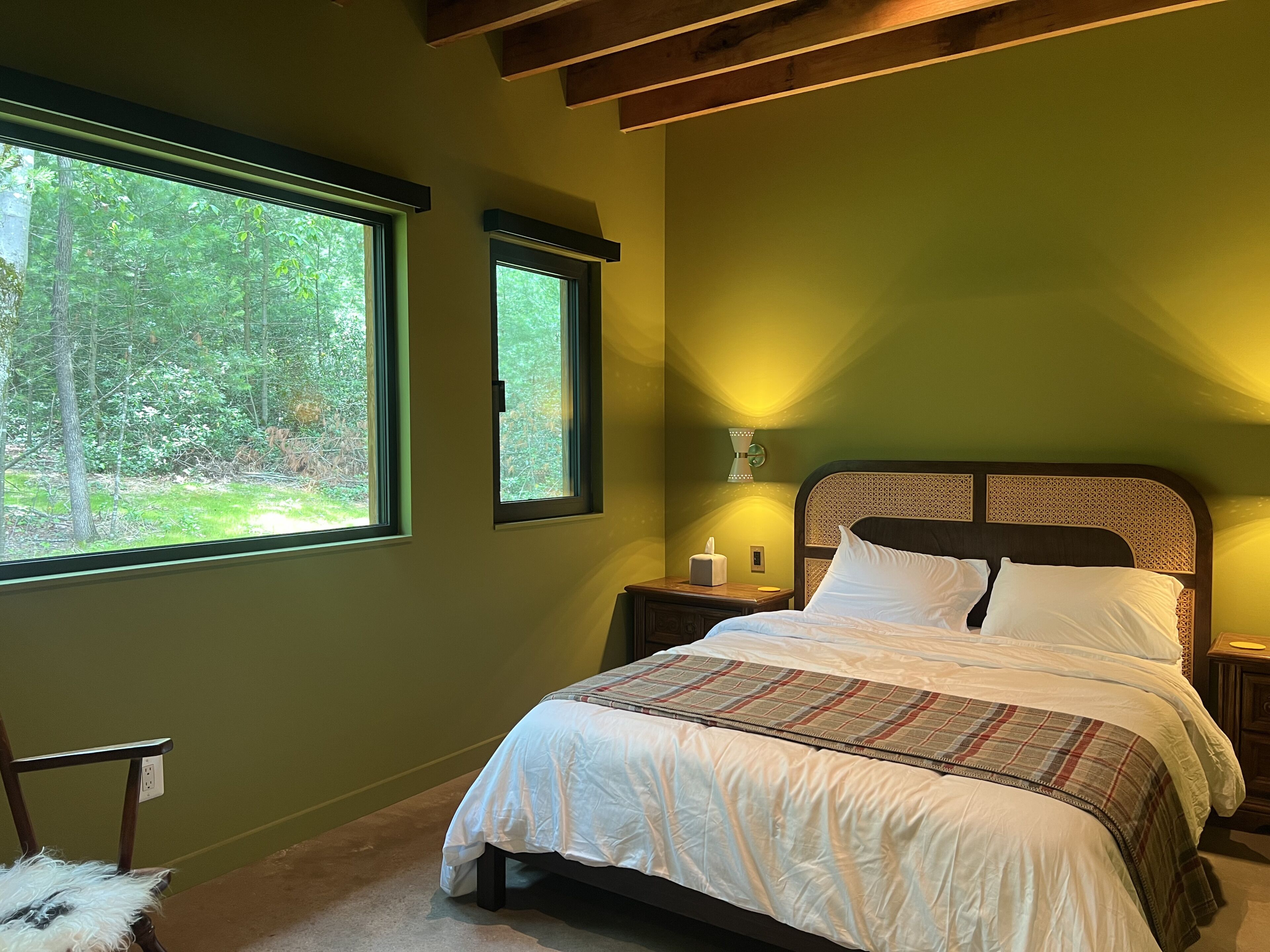 A bedroom with a large window, a bed with a plaid bedspread, a nightstand with a lamp, and a chair with a furry seat.