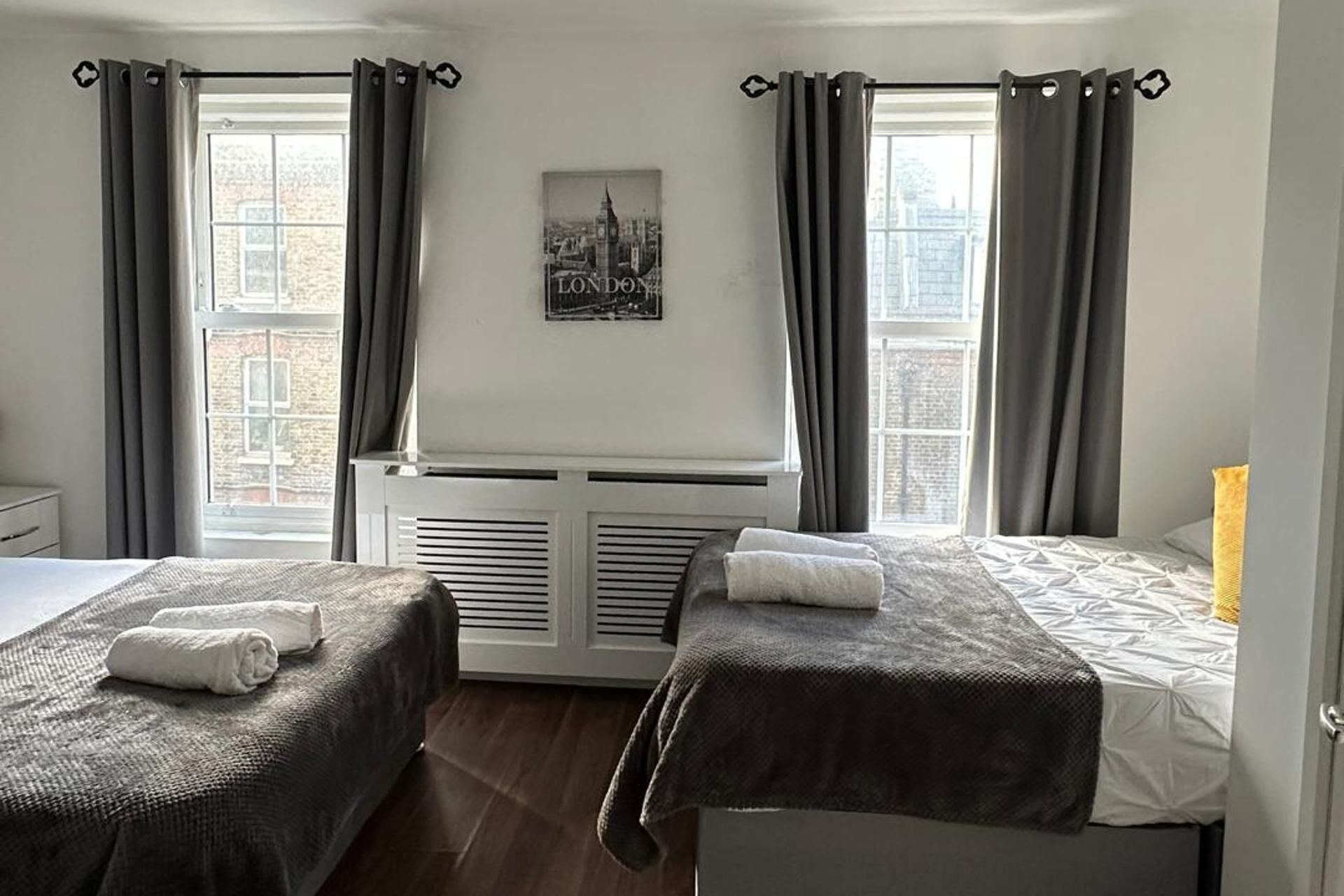 Ein Hotelzimmer mit zwei Betten, einem Heizkörper, einem Fenster mit Vorhängen und einem Stadtbild-Poster an der Wand.