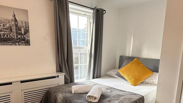 A hotel room with a bed, a window with curtains, a radiator, and a wall-mounted picture. - House Sleeps 22 With Wifi (London)